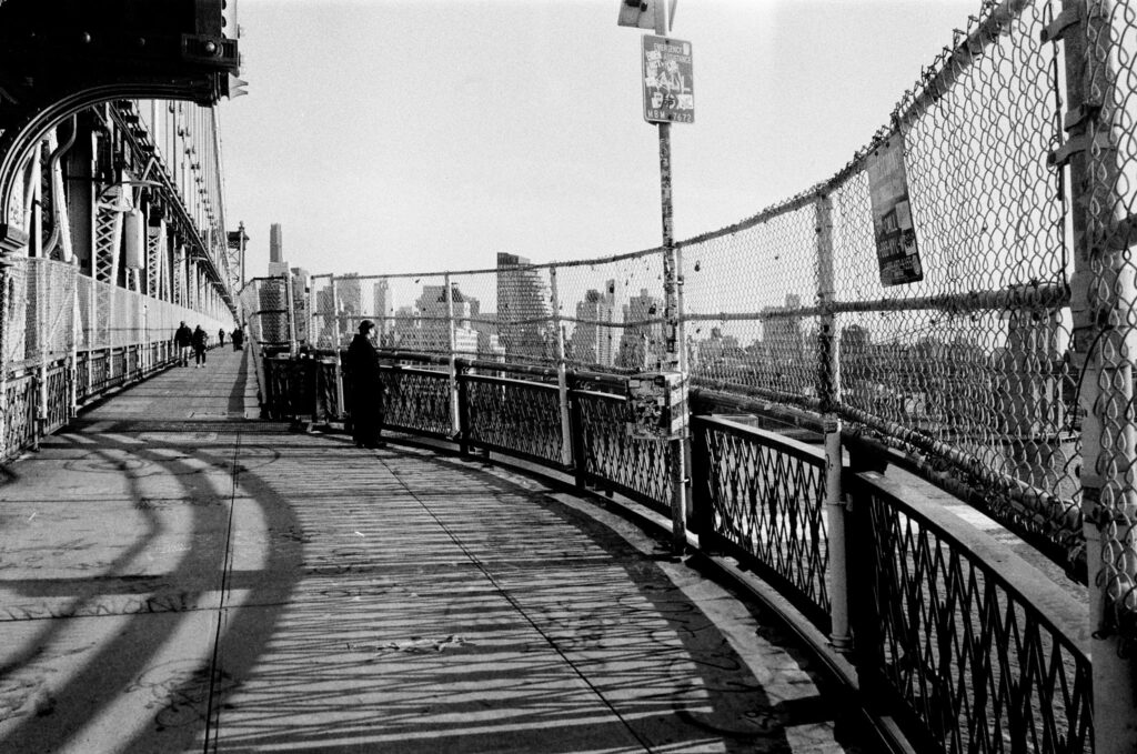 A black and white street photograph taken on the pedestrian walkway of the Williamsburg Bridge in New York City, looking toward Brooklyn in March 2025. The image features a curved chain-link fence on the right, covered in small locks and stickers, with the Brooklyn skyline visible in the distance. A person in dark clothing stands looking out over the water, while other pedestrians walk further down the path. Strong sunlight casts intricate, geometric shadows from the bridge structure across the walkway.